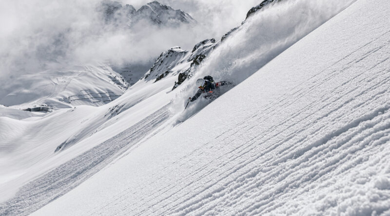 Ein Skifahrer fährt einen steilen, schneebedeckten Hang der Silvretta Montafon hinunter und wirbelt dabei Pulverschnee auf, während im Hintergrund schroffe Gipfel und Wolken zu sehen sind.