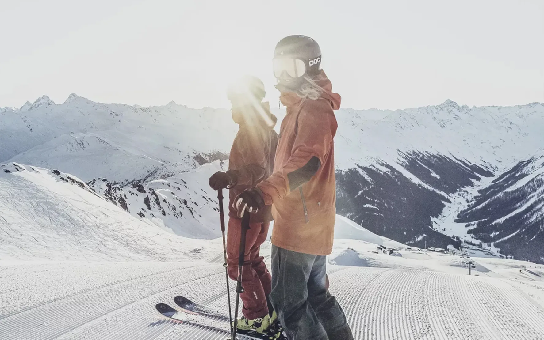 Two skiers in helmets and goggles stand on a snowy mountain slope at sunrise in Davos, with snow-covered peaks visible in the background.