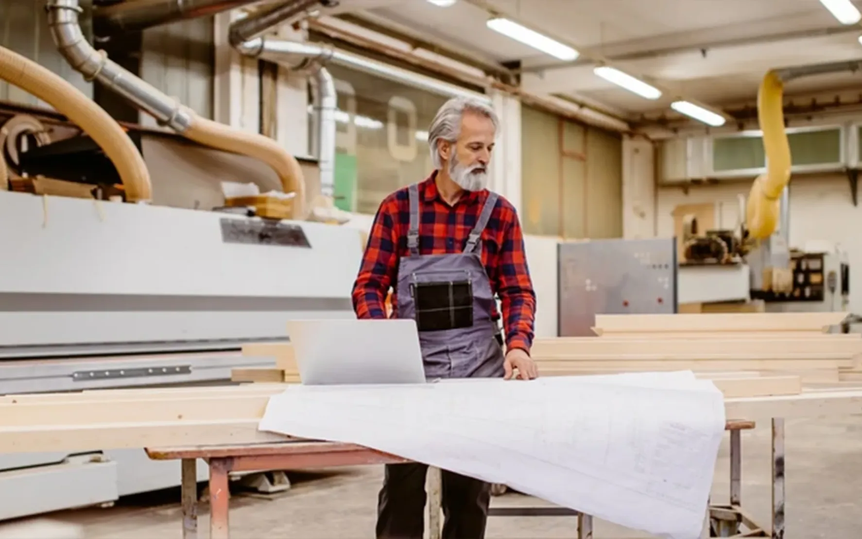 Handwerker mit grauem Bart betrachtet Baupläne in einer Werkstatt.