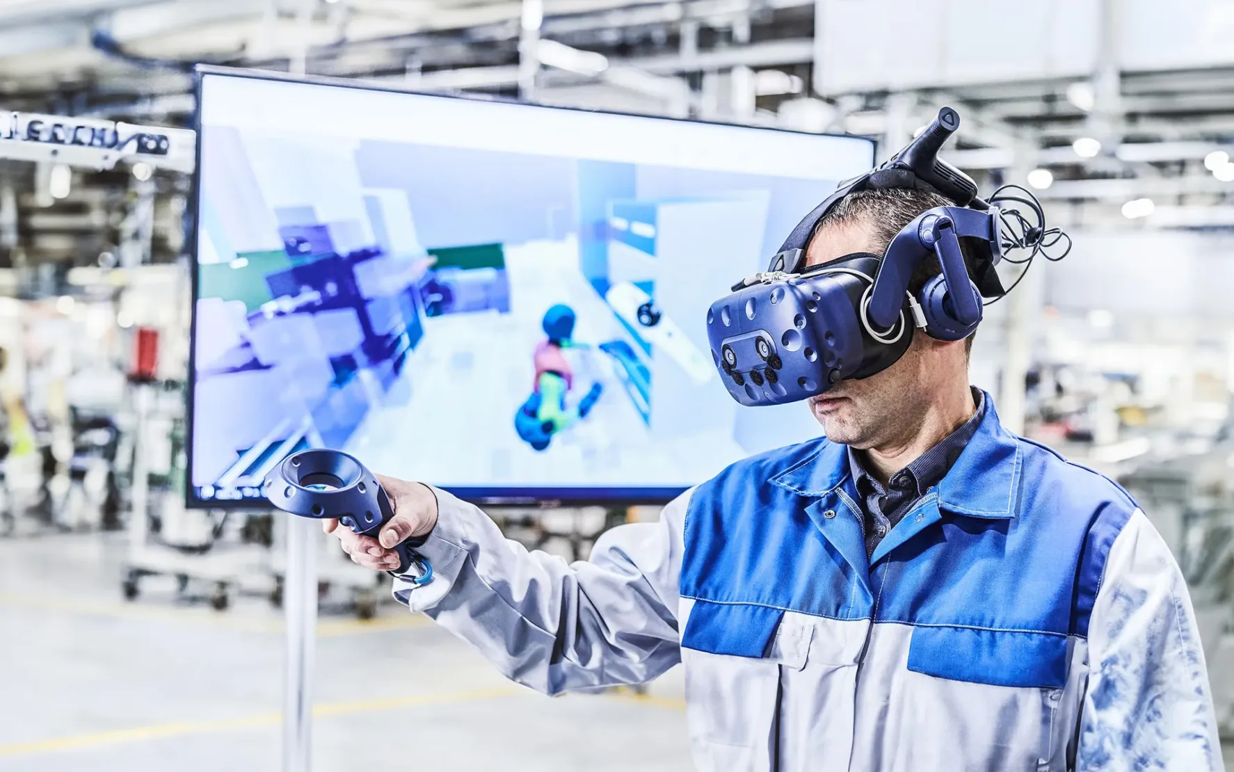 A man in a blue-grey work jacket is wearing VR goggles and holding a remote control. In the background, you can see a blurred screen showing the VR program.