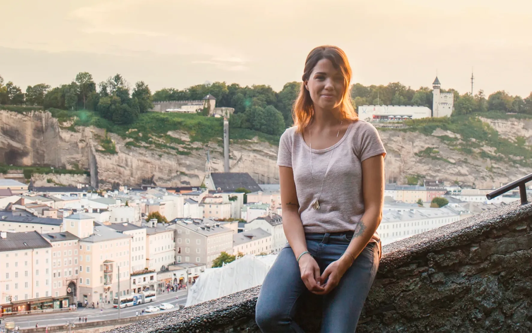 Eine Frau sitzt auf einer Mauer und lächelt in die Kamera. Im Hintergrund ist die Salzburger Altstadt mit Blick auf die das Museum der Moderne.