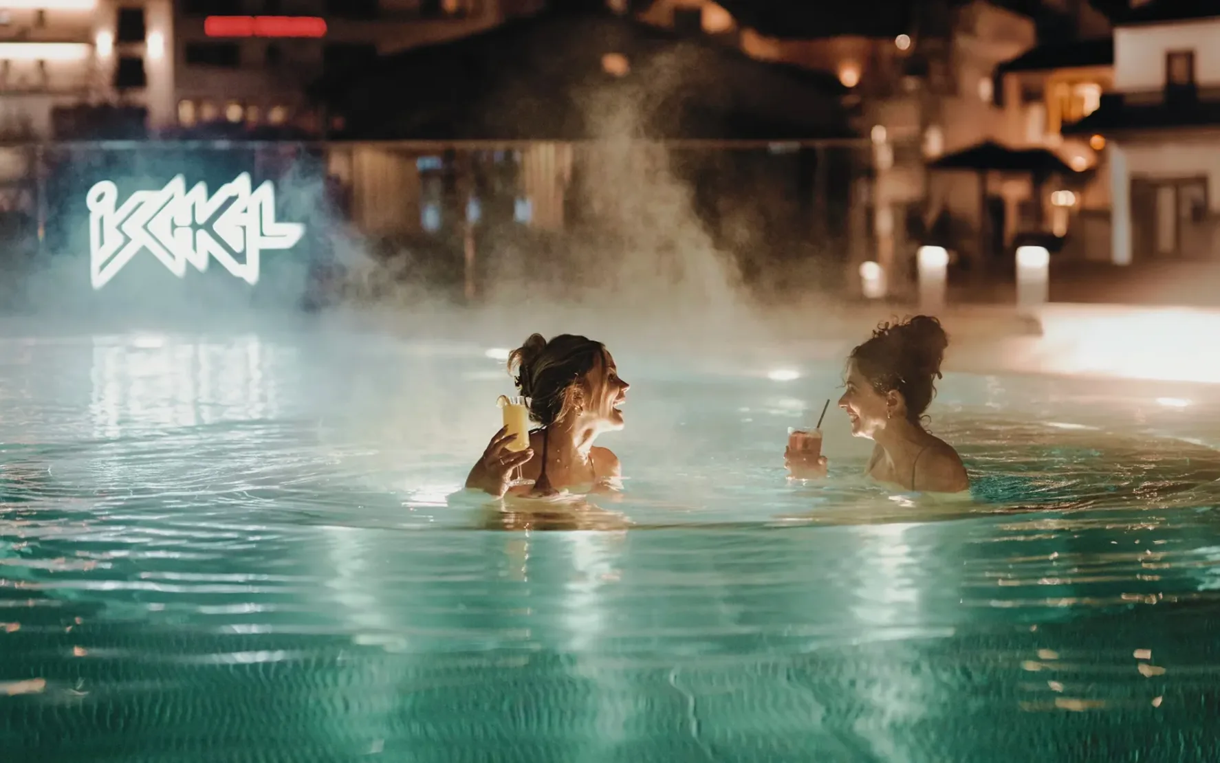 Zwei lachende Frauen genießen nachts mit Cocktails in der Hand das warme Außenbecken der Silvretta Therme Ischgl, umgeben von stimmungsvoll beleuchteten Gebäuden.