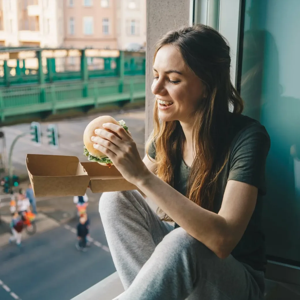 Woman eating take out burger at home in front of the open window