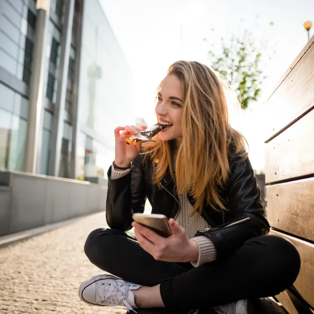 Woman eating chocolate on phone outdoor in street sitting on bench