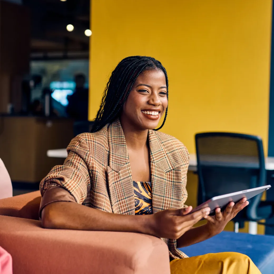 Eine Frau sitzt auf einer Couch, hält ein Tablet in der Hand und lächelt. Neben ihr sitzt ein Mann in einer modernen, farbenfrohen Büroumgebung, die ihr Engagement für nachhaltige Beschaffung widerspiegelt.