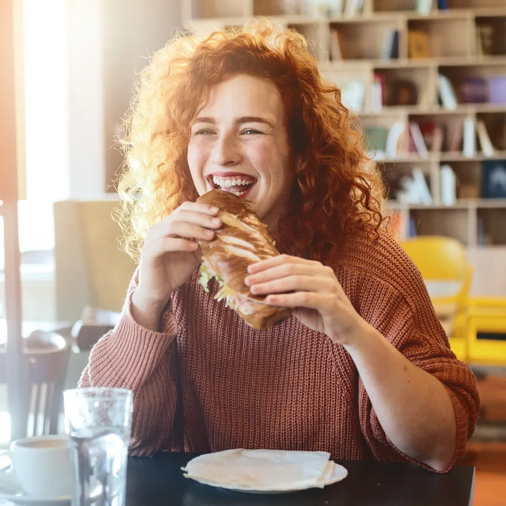 Frau mit lockigem rotem Haar, die einen rosa Pullover trägt, lächelt, während sie nach einem optimierten Einkauf ein Sandwich in einem Café genießt. Ein Glas Wasser und eine Serviette stehen auf dem Tisch, im Hintergrund stehen Bücherregale und Stühle.