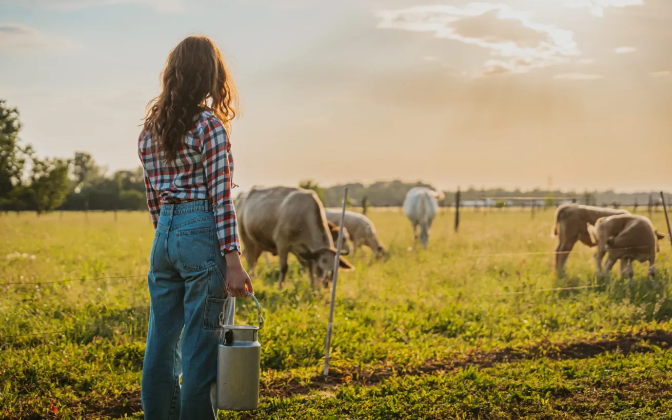 Young woman holding milk canister while watching over cows grazing on field